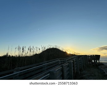 A scenic view of a beach at sunrise with a wooden boardwalk leading towards the beach. Tall grasses and sand dunes frame the path, while the sun casts a warm glow over the horizon. - Powered by Shutterstock