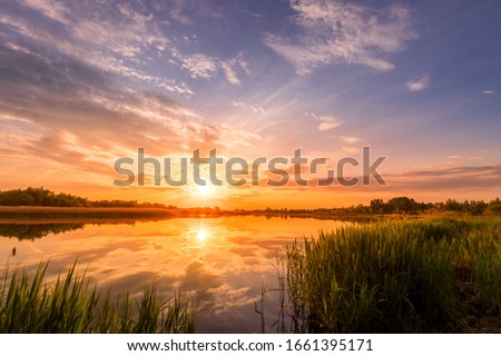 Scenic view of beautiful sunrise or dawn above the pond or lake at spring or early summer morning with cloudy sky background, fog over water and reed grass with dew at foreground. Water reflection.