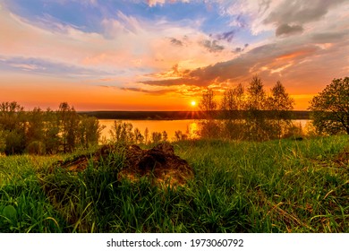 Scenic View At Beautiful Sunset On A Shiny Lake With Old Rough Stone On The Foreground, Green Grass, Birch Trees, Golden Sun Rays, Calm Water ,nice Cloudy Sky On A Background, Spring Landscape