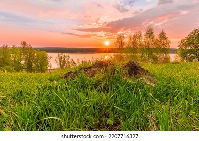 Scenic View At Beautiful Sunset On A Shiny Lake With Old Rough Stone On The Foreground, Green Grass, Birch Trees, Golden Sun Rays, Calm Water ,nice Cloudy Sky On A Background, Spring Landscape