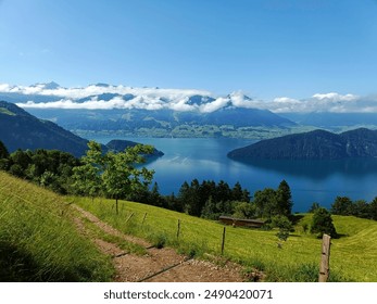 A scenic view of a blue lake surrounded by green hills and mountains under a bright blue sky with scattered clouds. A dirt path leads down the grassy slope, evoking peace and natural beauty. - Powered by Shutterstock
