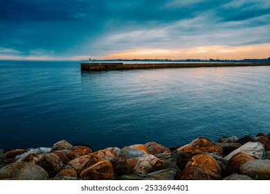 Scenic view of a calm sea with a rocky shore and a distant pier under a dramatic cloudy sky at sunset. - Powered by Shutterstock