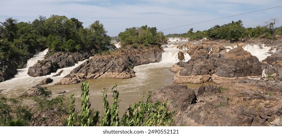 Scenic view of cascading waterfalls surrounded by rocky terrain and lush greenery under a partly cloudy sky. in Laos - Powered by Shutterstock