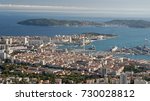 A scenic view of the city of Toulon from a hill called "Mount Faron". Houses and buildings have red tiled roofs and big ships are parked in the harbor. Two islands are visible in the background.