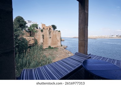 A scenic view of a coastal area with a historic fort wall, lush greenery, and a river. A terrace with blue tables and chairs, enjoying the view under a partly cloudy sky.
