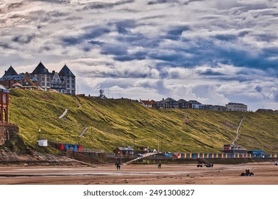 A scenic view of a coastal town with a grassy hill, colorful beach huts, and people walking on the sandy beach under a cloudy sky. - Powered by Shutterstock