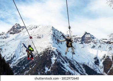 Scenic View Couple Of Tourist With Skiing Equipment Enjoy Having Fun Riding Extreme Suspended Zipline Wire Flying Over Gorge Canyon Against Snowcapped Mountains On Background. Adrenaline Recreation