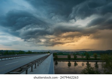 A scenic view of dark clouds loom above a bridge over a river, surrounded by trees - Powered by Shutterstock