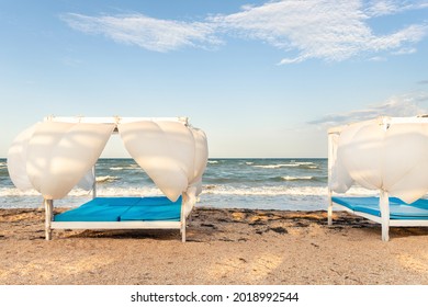 Scenic View Empty White Beach Wooden Canopies Cabanas With White Curtain Blowing By Wind Against Stormy Sea Or Ocean San Seaside. Luxury Sunbed For Spa And Leisure Vacation Travel