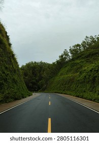 Scenic view of an empty winding asphalt road cutting through lush green hills and forest under a cloudy overcast sky.