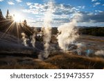 A scenic view of a geothermal landscape filled with numerous steam vents, with the sun shining through trees on the left, casting a radiant glow. The sky is partly cloudy, and the background features