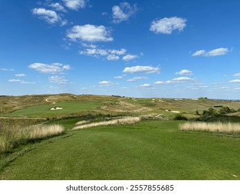 A scenic view of a golf course on a sunny day, featuring lush green fairways, rolling hills, and patches of tall golden grasses.  - Powered by Shutterstock