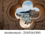 A scenic view of Dubrovnik’s harbor framed through a decorative stone window, capturing boats, fort walls, and the Adriatic Sea. Perfect for travel, architecture, and historic city themes.