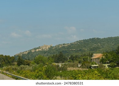 A scenic view of a hilltop village, nestled amongst lush greenery and trees, with a paved road winding through the foreground. - Powered by Shutterstock