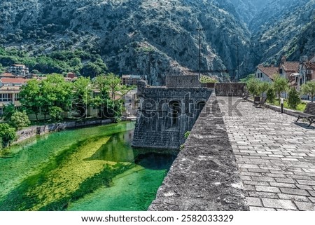Scenic view of the historic Kotor city walls, stretching along a green canal with the majestic Montenegrin mountains in the background. The blend of nature and medieval architecture