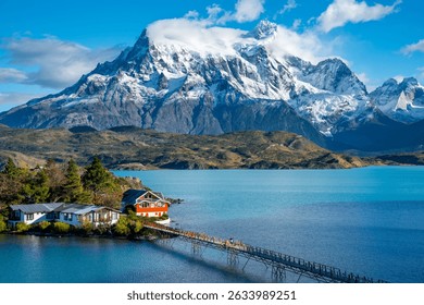 Scenic view of Lake Nordenskjöld in Chilean Patagonia, with snowy mountains in the background and a small lodge connected by a bridge in the foreground.