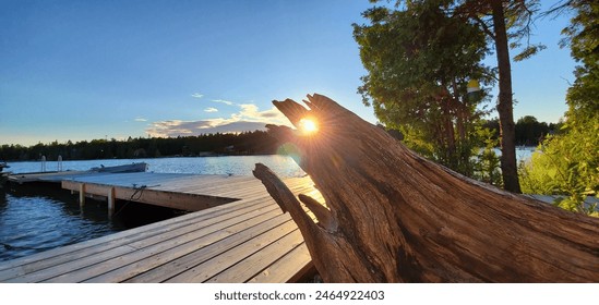 A scenic view of a lakeside wooden dock at sunset, with the sun peeking through a weathered tree stump and lush greenery in the background. - Powered by Shutterstock