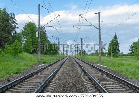 A scenic view looking down a long stretch of electrified double railway tracks cutting through a lush green landscape under a bright, cloudy sky. This expansive perspective captures the mesmerizing symmetry of parallel steel rails leading toward the distant horizon, flanked by vibrant grass and dense woodland. The overhead catenary wires, supported by sturdy concrete poles, create intricate geometric patterns against the soft daylight. The ballast stones between the sleepers add textured detail to the foreground, while the gentle curve of the scenery suggests a quiet, peaceful journey through the countryside. Nature and industrial infrastructure blend seamlessly, illustrating a vital transport artery that connects remote regions. The composition emphasizes depth, inviting the viewer to imagine traveling along this path, with the vast horizon line drawing the eye further into the frame. The overcast sky softens the natural colors, creating a serene and timeless atmosphere typical of rural train routes. It is a striking representation of modern transit architecture integrated into the environment, offering a sense of stability, travel, and exploration. The absence of trains allows the focus to remain on the clean lines and the stark geometry of the track system, highlighting both engineering precision and the beauty of the surrounding landscape. The quiet stillness suggests a moment of pause before the next transit activity, making the image ideal for themes of logistics, journey, infrastructure, and rural development. Every element, from the weathered poles to the carefully arranged gravel, contributes to a balanced, professional shot that captures the essence of rail travel.