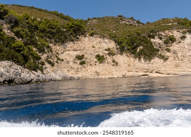 A scenic view of lush green hills and limestone cliffs meeting the calm waters along Zakynthos Island’s coastline, Greece, with waves from a passing boat in the foreground. - Powered by Shutterstock