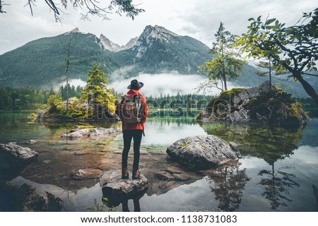 Scenic view of a man wearing a red jacket, a hat and a backpack looking at mountains, Lake Hintersee, Berchtesgaden, Germany 