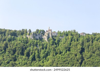 A scenic view of a medieval castle perched on a rocky hilltop, surrounded by lush green forests under a clear blue sky. - Powered by Shutterstock