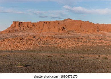 Scenic view of the Mongolian Flaming Cliffs tourist attraction in the Gobi Desert of Mongolia at sunset with distant tourists on the red rocks of the ridge and their tourist vehicle at the bottom 