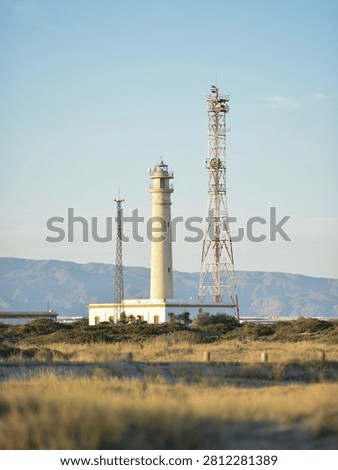 Scenic view of an old functional lighthouse and a tall industrial radio antenna tower, situated in a coastal area with dry grass and distant mountains in the background.