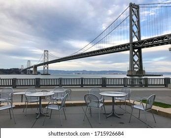 Scenic View On San Francisco – Oakland Bay Bridge In California, USA With Empty Cafe Tables And Chairs Foreground