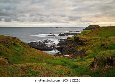 A scenic view overlooking green grassy hills with beautiful volcanic, rocky coastline in the background, deep blue sea and cloudy sky on the horizon, at Phillip Island in Australia.