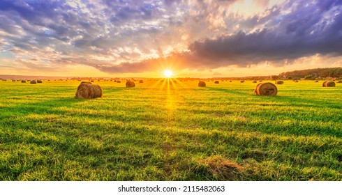 Scenic view at picturesque burning sunset in a green shiny field with hay stacks, bright cloudy sky , golden sun rays and road leading far away, summer valley landscape