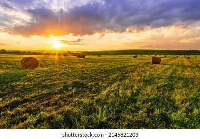 Scenic view at picturesque burning sunset in a green shiny field with hay stacks, bright cloudy sky , trees and golden sun rays, summer valley landscape