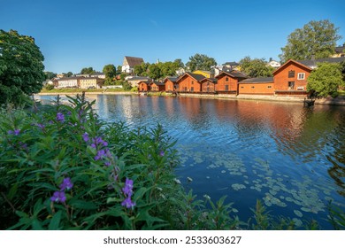 A scenic view of Porvoo, Finland, with traditional red wooden riverside houses, a clear blue sky, and vibrant green foliage in the foreground. - Powered by Shutterstock