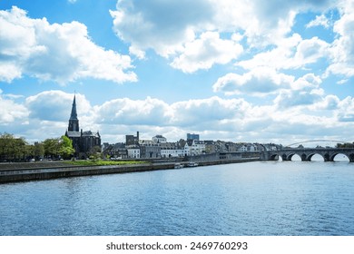 Scenic view of a quaint European town Maastricht with a historic church spire overlooking Meuse river and a stone bridge under a partly cloudy sky - Powered by Shutterstock