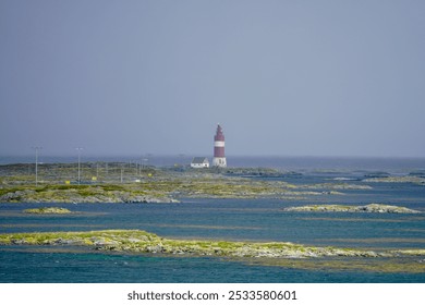 A scenic view of a red and white lighthouse on a rocky island surrounded by blue ocean under a clear sky. - Powered by Shutterstock