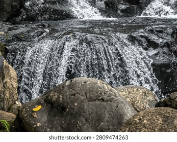 Scenic view of a refreshing waterfall cascade flowing over black volcanic rocks behind large natural river stones in the wild jungle.