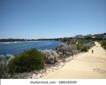 A Scenic View Of Rocky Bay In The Swan River Perth. Two Dogs Walking Down A Riverside Path On A Sunny Day. Boats Traveling Downstream Leaving A Wake Between The Channel Markers.