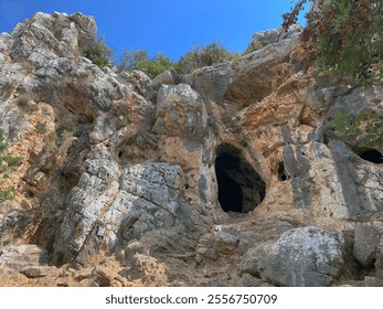 A scenic view of a rocky cliff with natural caves and rugged formations under a clear blue sky. - Powered by Shutterstock