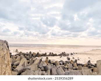 Scenic view of a rocky shoreline with concrete breakwaters, capturing the dynamic meeting of ocean waves and sky under a cloudy sunset. Perfect for coastal themes and nature-inspired projects. - Powered by Shutterstock