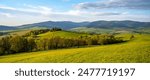 A scenic view of rolling green hills and distant mountains in the Kralicky Sneznik Mountains of Czechia. The landscape is bathed in warm sunlight, creating a serene and peaceful atmosphere.