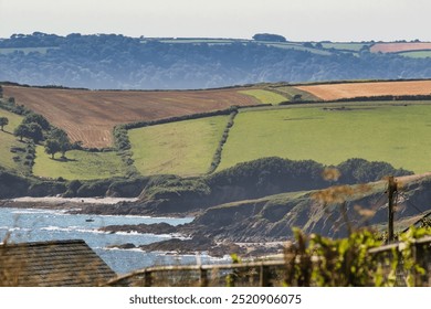 A scenic view of rolling green hills and fields by the coast, with a clear blue sea and rocky shoreline. Cornwall, UK - Powered by Shutterstock