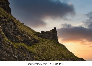 A scenic view of a rugged cliff with remnants of an ancient stone structure at sunset. The sky is filled with dramatic clouds, casting a warm glow over the landscape.At Tintagel, Cornwall, UK. - Powered by Shutterstock