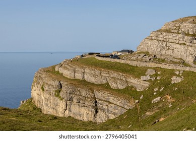 A scenic view of a rugged cliffside path overlooking the sea, with a house and vehicles in the distance at Great Orme, in Llandudno, North Wales.