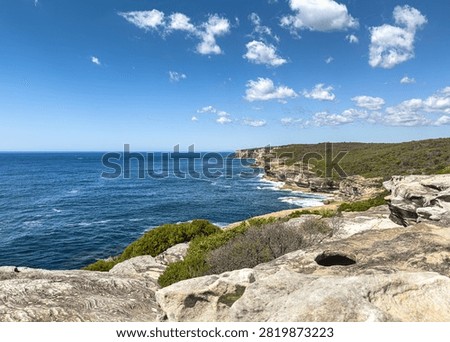 Scenic view of sandstone cliffs overlooking the deep blue ocean along the Australian coastline under a bright sky with scattered clouds. A dramatic coastal landscape featuring rugged rock formations, native vegetation, and expansive sea views.