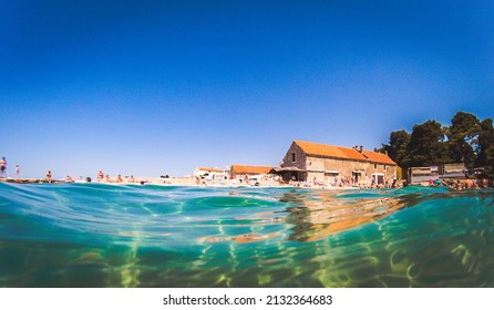 A Scenic View Of A Seascape With A Background Of People Sunbathing Against Old Stone Buildings On A Sunny Day