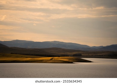 A scenic view of a serene lake with rolling hills and mountains under a cloudy sky at sunset, in Nei Mongol, China. - Powered by Shutterstock