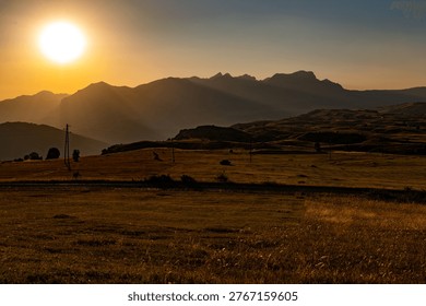 Scenic view of a setting sun casting warm rays over silhouetted mountains and rolling hills, with a vast field in the foreground