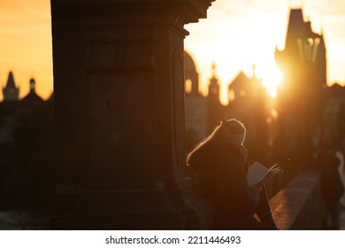 Scenic View With A Silhouette Of A Young Woman Standing With The Back And Reading On Charles Bridge From Prague During An Amazing Autumn Sunrise. Reading In Landmark Places Around The World.