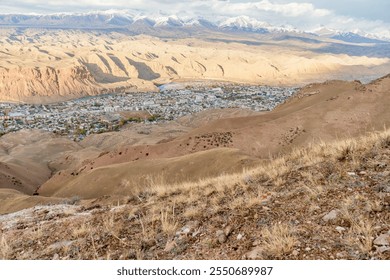 A scenic view of a small town nestled among arid hills and dramatic canyon landscapes. Snow-capped mountains rise in the background, contrasting with the desert terrain under a partially cloudy sky. - Powered by Shutterstock