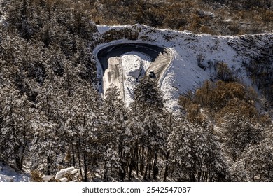 Scenic view of snow-covered mountains. Snow blankets the trees and hillsides, creating a serene winter landscape under bright sunlight. The road curves along the hillside, framed by evergreen trees. - Powered by Shutterstock