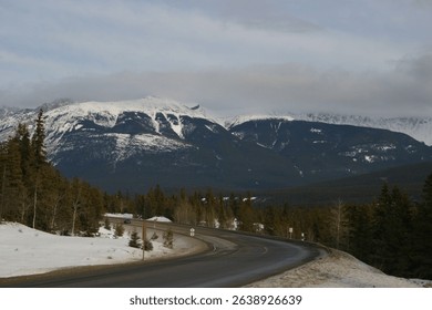 Scenic view of a snowy mountain range with a winding road and evergreen trees under a cloudy sky, perfect for nature and travel themes.